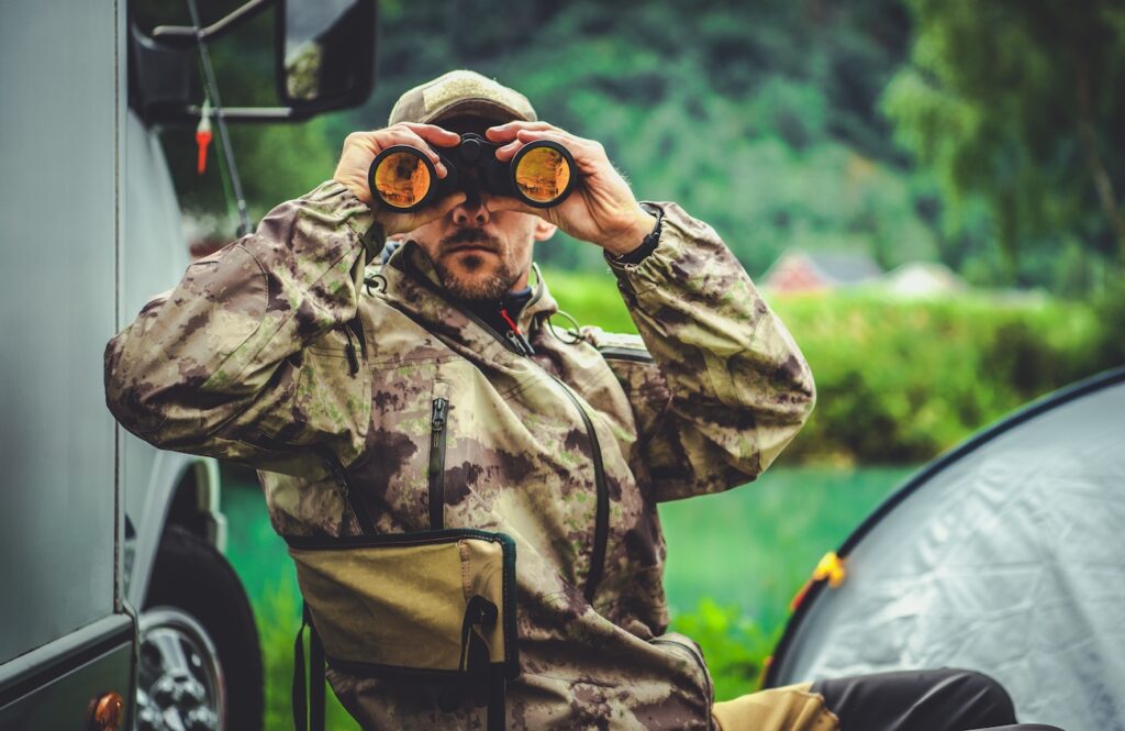 Hunting Season Game Spotting. Caucasian Hunter with Binoculars and the Campsite in Background.